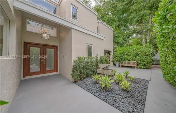 Stucco exterior featuring double glass entry doors, geometric star pendant lighting, expansive concrete patio flooring, and a stone-bordered garden bed with drought-tolerant plantings