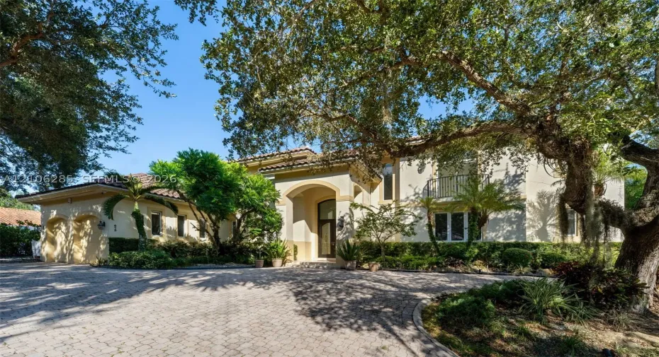 Stucco exterior featuring an arched entryway, tile roof, multi-car garage, upper-level balcony with metal railing, and paver driveway