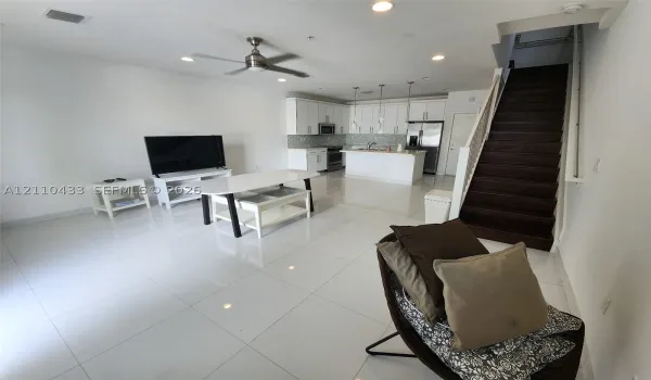 Open-concept interior featuring large-format tile flooring, white cabinetry, a kitchen island, recessed lighting, and a staircase with dark wood-finish treads