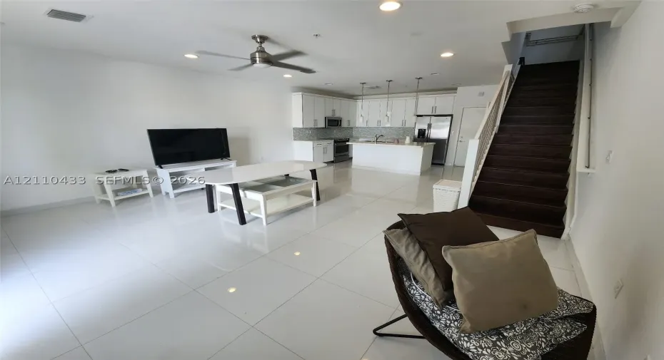 Open-concept interior featuring large-format tile flooring, white cabinetry, a kitchen island, recessed lighting, and a staircase with dark wood-finish treads