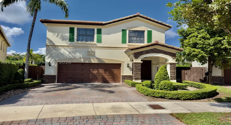 Stucco exterior featuring decorative green shutters, stone-veneer accents, a paved driveway, and manicured landscaping