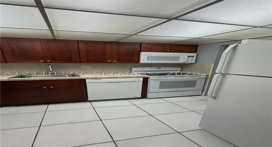 Kitchen featuring stained wood cabinetry, tile countertops, integrated white appliances, and a grid-panel drop ceiling