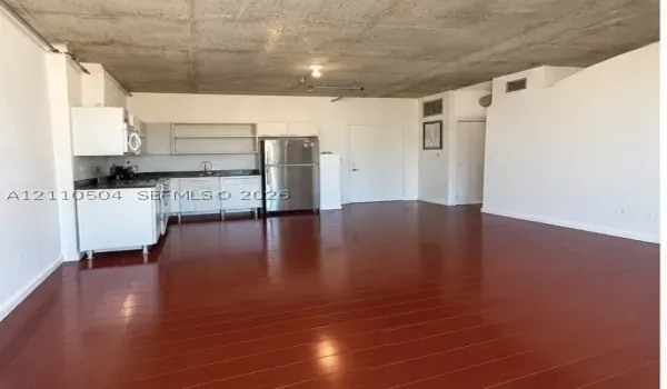 Industrial loft interior featuring exposed raw concrete ceilings, wood-finish flooring, white appliances, and white cabinetry with dark countertops