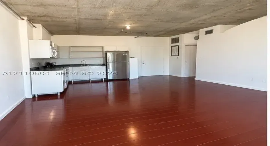 Industrial loft interior featuring exposed raw concrete ceilings, wood-finish flooring, white appliances, and white cabinetry with dark countertops