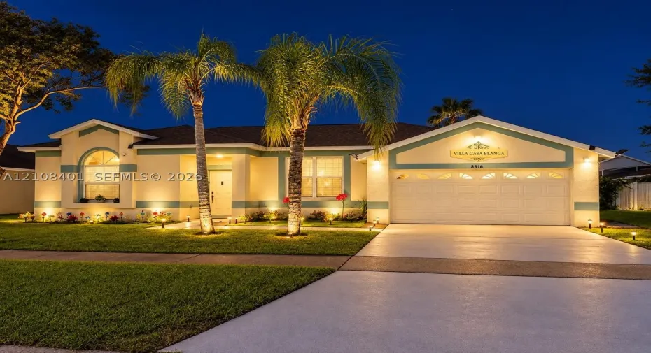 Single-story exterior featuring a stucco facade, arched window detail, two-car garage, concrete driveway, and mature palm landscaping