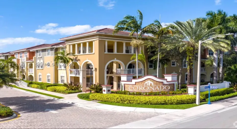 Mediterranean-style multi-story building featuring arched architectural elements, recessed balconies with metal railings, a tiled roof, and professional landscaping with mature palm trees