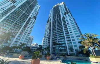 High-rise residential tower featuring glass-panel balconies, concrete facade, exterior terrace, and ground-level pool area with palm tree landscaping