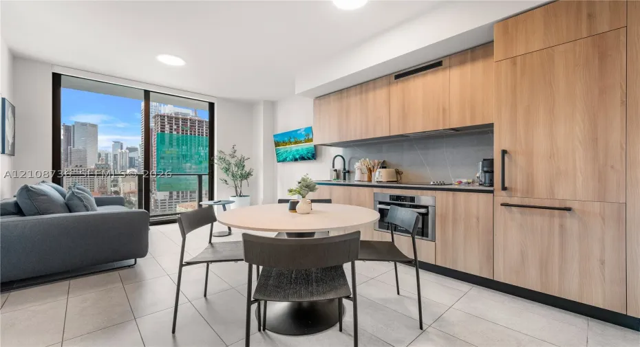 Contemporary kitchen featuring light wood-finish cabinetry, integrated panel-ready appliances, stone-composite backsplash, floor-to-ceiling glass sliding door, and neutral-toned tile flooring