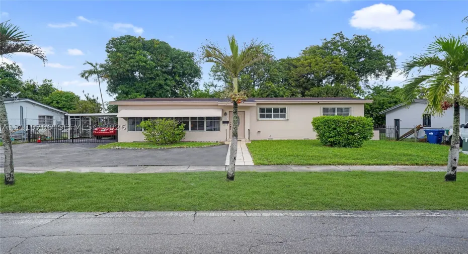 Single-story exterior featuring a light-toned masonry facade, asphalt driveway, covered metal carport, and manicured landscaping