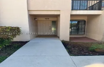 Exterior entryway featuring a concrete walkway, stucco facade, paneled door with a sidelight, and an upper-level balcony with metal railing