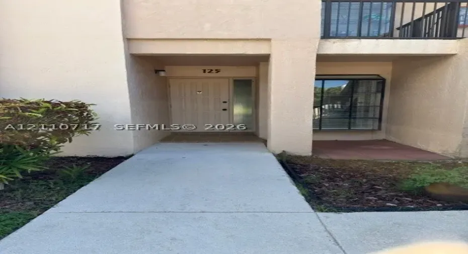 Exterior entryway featuring a concrete walkway, stucco facade, paneled door with a sidelight, and an upper-level balcony with metal railing