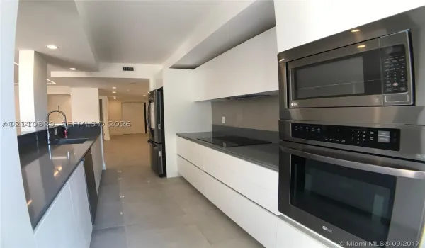 Kitchen featuring white flat-panel cabinetry, charcoal solid-surface countertops, stainless steel built-in double oven and microwave, recessed lighting, and large-format tile flooring