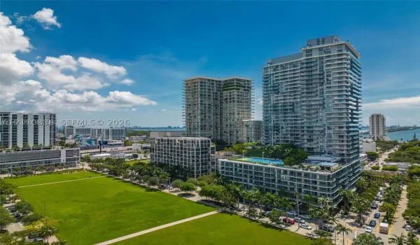 High-rise condominium towers featuring glass-paneled balconies and a multi-level recreational deck with a swimming pool, situated beside a expansive lawn