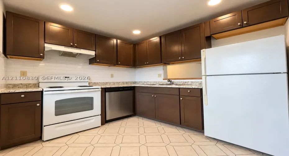Spacious kitchen featuring rich wood cabinetry with ample storage, granite-style countertops, stainless steel appliances including a range and dishwasher, white refrigerator, and ceramic tile flooring throughout