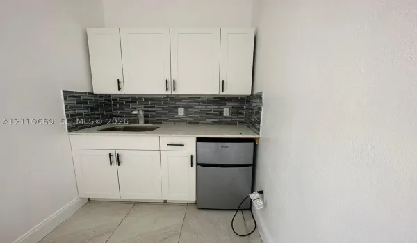 Kitchenette featuring white cabinetry with matte black hardware, stone-finish countertop, stainless steel sink with high-arc faucet, linear mosaic tile backsplash, and built-in under-counter refrigerator