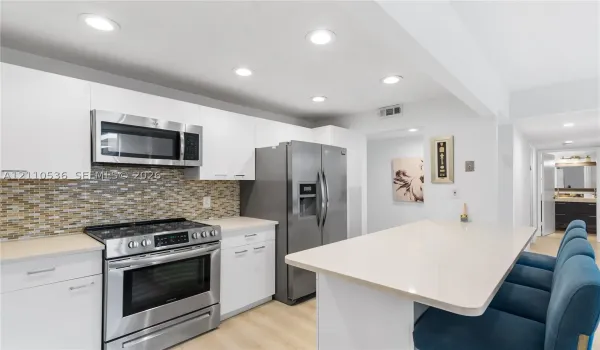 Kitchen featuring flat-panel white cabinetry, stainless steel appliances, multi-tonal glass tile backsplash, light-toned countertop, and recessed lighting