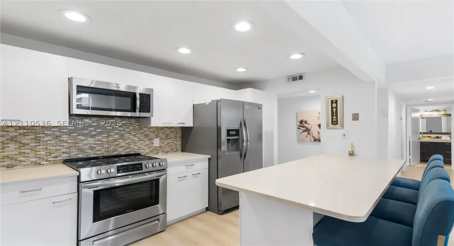 Kitchen featuring flat-panel white cabinetry, stainless steel appliances, multi-tonal glass tile backsplash, light-toned countertop, and recessed lighting