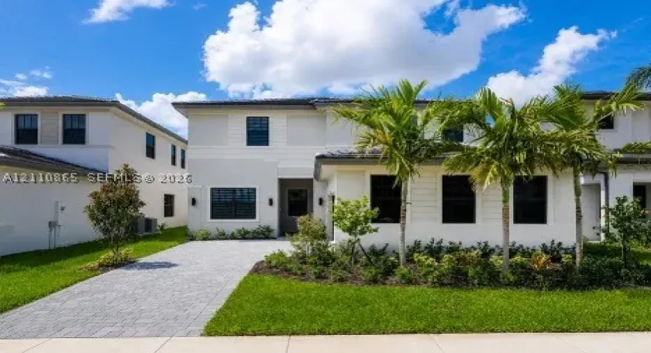 Two-story exterior featuring light-toned smooth stucco finish, horizontal siding accents, dark-framed windows, paver driveway, and professional landscaping with palm trees
