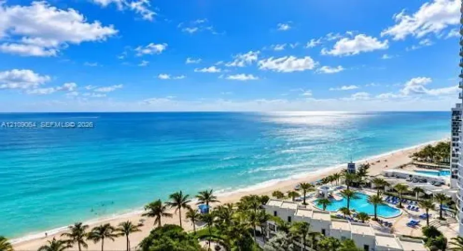 High-altitude perspective featuring an expansive ocean horizon, sandy coastline, and a multi-pool aquatic complex with surrounding palm trees