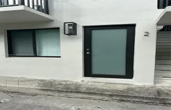 Exterior entrance featuring a white stucco facade, a black-framed door with a frosted glass panel, a sleek rectangular wall-mounted light fixture, and a black-framed window