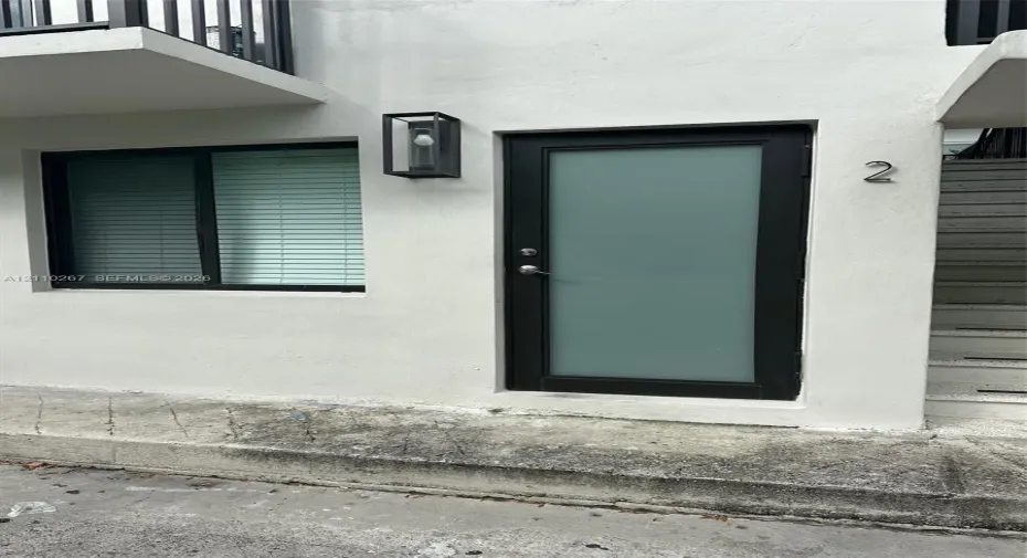 Exterior entrance featuring a white stucco facade, a black-framed door with a frosted glass panel, a sleek rectangular wall-mounted light fixture, and a black-framed window