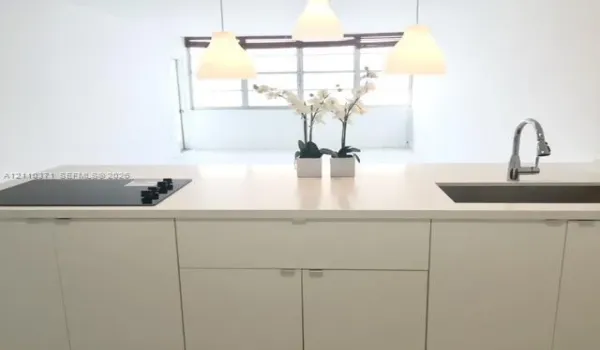 Kitchen island featuring a solid-surface countertop, integrated flat-surface cooktop, undermount stainless steel sink with a high-arch gooseneck faucet, minimalist flat-panel cabinetry, and three pendant lights