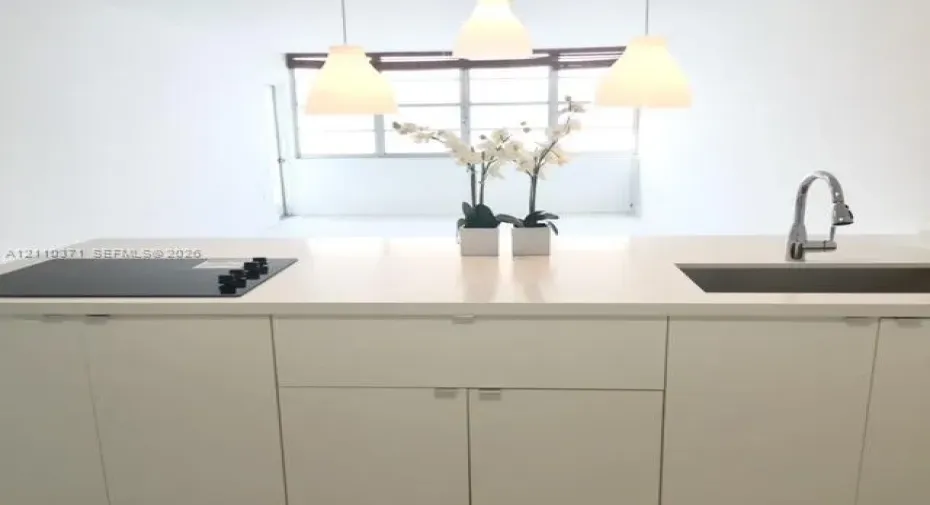Kitchen island featuring a solid-surface countertop, integrated flat-surface cooktop, undermount stainless steel sink with a high-arch gooseneck faucet, minimalist flat-panel cabinetry, and three pendant lights