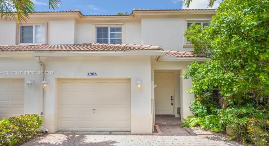 Two-story stucco exterior featuring a tiled roof, attached single-car garage, brick paver driveway, and wall-mounted exterior light fixtures