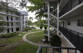 Multi-story building exterior featuring stucco siding, black metal balcony railings, open-air staircases, and a paved walkway through landscaped grounds