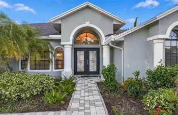 Stucco exterior featuring dual columns, a stone paver walkway, double front entry doors with decorative glass inserts, and a prominent arched transom window