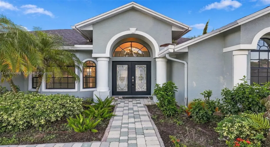 Stucco exterior featuring dual columns, a stone paver walkway, double front entry doors with decorative glass inserts, and a prominent arched transom window