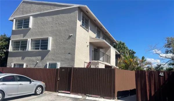 Multi-level building exterior with textured stucco siding, horizontal sliding windows, a private second-story balcony with metal railing, and a perimeter corrugated metal fence