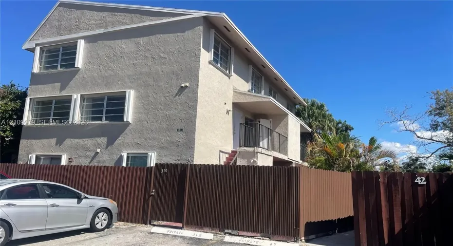 Multi-level building exterior with textured stucco siding, horizontal sliding windows, a private second-story balcony with metal railing, and a perimeter corrugated metal fence