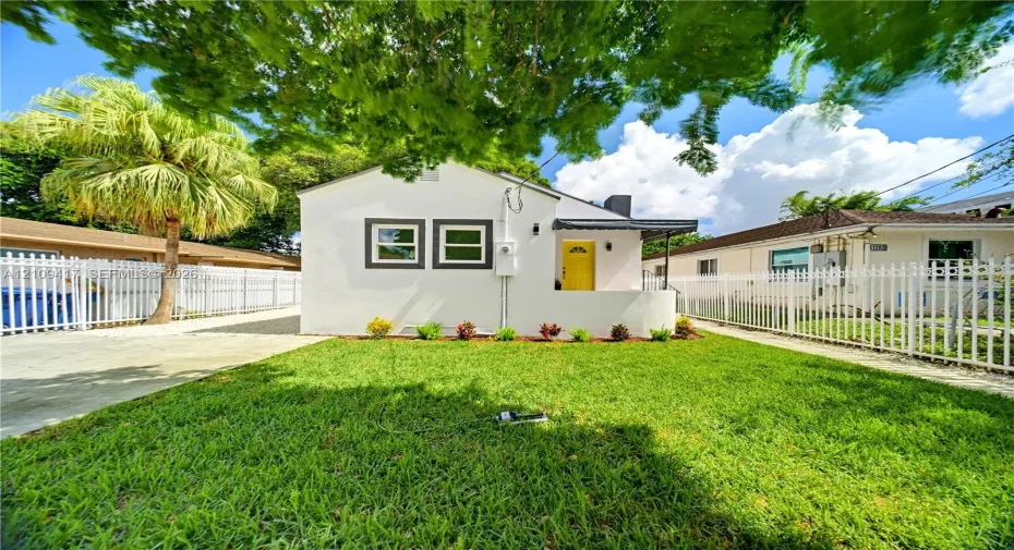 Single-story exterior featuring a light-toned stucco facade, a vibrant front entry door, a covered porch, and perimeter white metal fencing