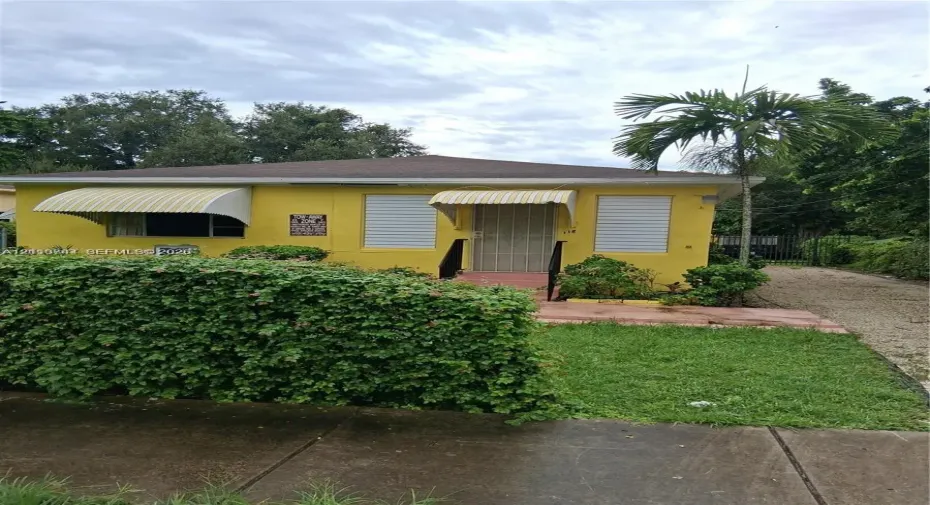 Single-story residence featuring a stucco exterior, metal window awnings, fixed metal security shutters, and a gravel driveway