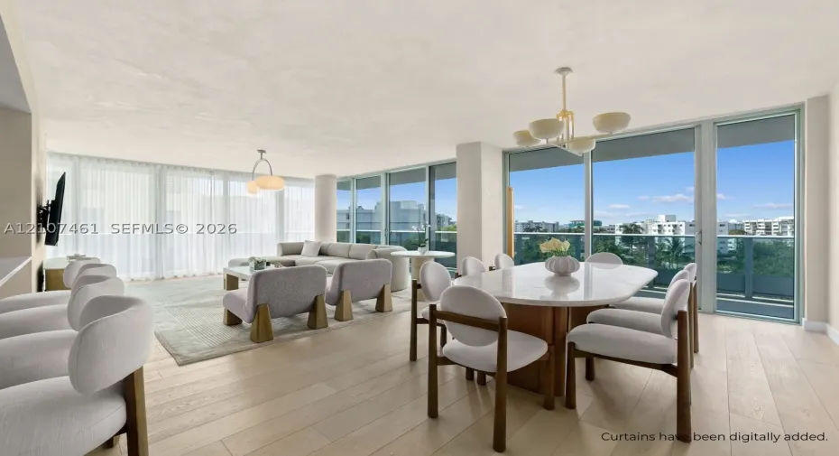 Open-concept area featuring light-toned wood-finish flooring, floor-to-ceiling glass paneling, metal-framed terrace access, and dual pendant chandeliers