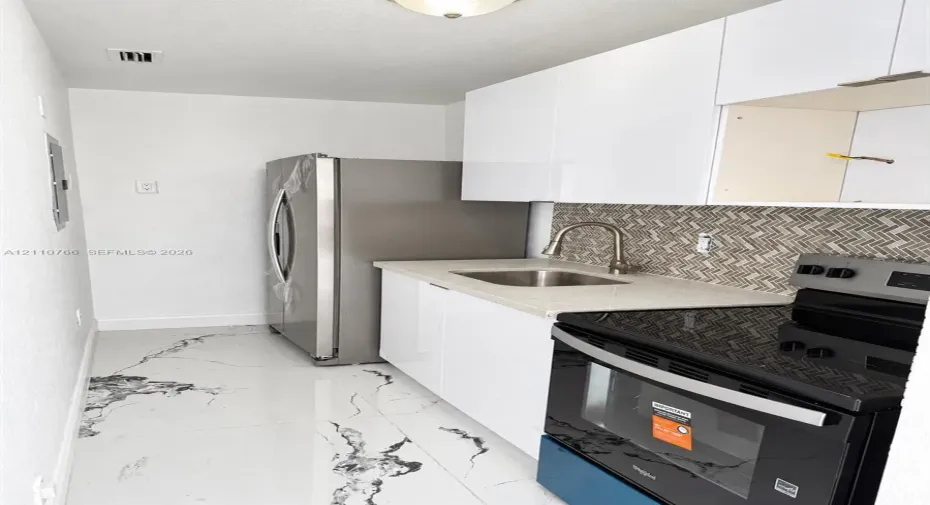 Kitchen featuring white gloss cabinetry, stone-finish countertops, stainless steel refrigerator, black finish range, and herringbone tile backsplash over marble-finish flooring