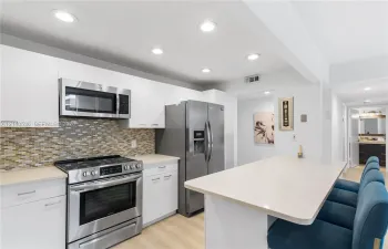 Kitchen featuring flat-panel white cabinetry, stainless steel appliances, multi-tonal glass tile backsplash, light-toned countertop, and recessed lighting
