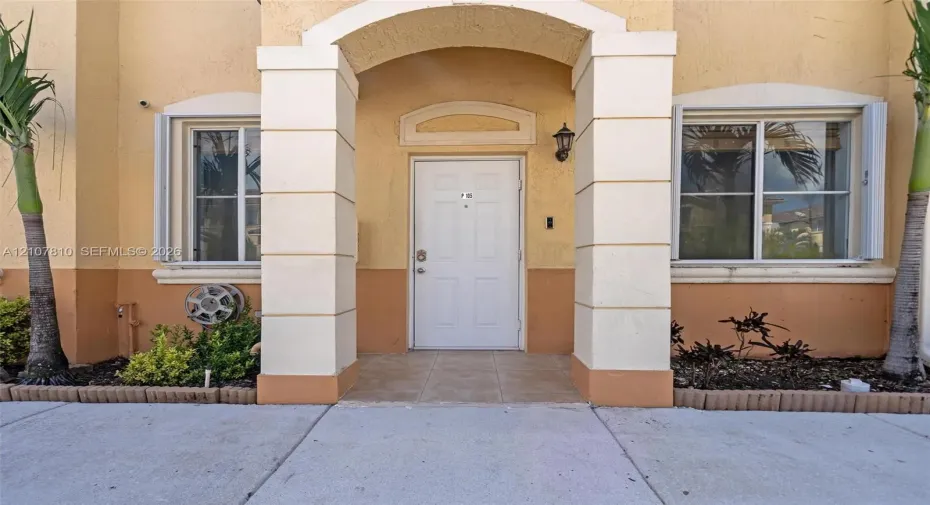 Stucco exterior entrance featuring a covered arched portico with decorative columns, tile porch flooring, and two dual-pane windows with integrated shutters