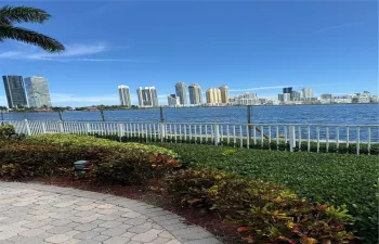 Waterfront property featuring a paved stone walkway, landscaped shrubbery, a white perimeter fence, and a skyline backdrop