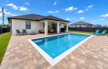 Stucco exterior with a covered patio, featuring an inground swimming pool surrounded by paved hardscaping and enclosed by a perimeter fence
