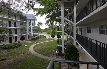Multi-story building exterior featuring stucco siding, black metal balcony railings, open-air staircases, and a paved walkway through landscaped grounds