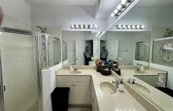 L-shaped vanity featuring dual integrated sinks, light-toned countertop, extensive wall-spanning mirror, and dual over-mirror light fixtures, complemented by a glass-enclosed shower stall