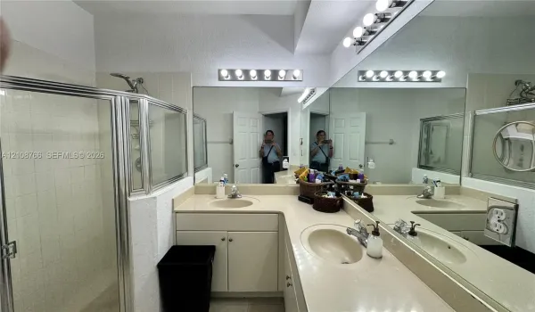 L-shaped vanity featuring dual integrated sinks, light-toned countertop, extensive wall-spanning mirror, and dual over-mirror light fixtures, complemented by a glass-enclosed shower stall