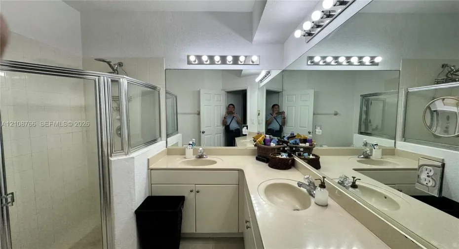 L-shaped vanity featuring dual integrated sinks, light-toned countertop, extensive wall-spanning mirror, and dual over-mirror light fixtures, complemented by a glass-enclosed shower stall