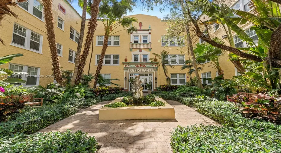 Stucco exterior featuring decorative masonry accents, a central courtyard fountain, multi-pane windows, and paved walkways surrounded by lush mature landscaping