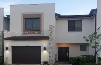 Modern stucco facade featuring horizontal wood-finish siding accents, decorative stone veneer pillars, integrated wall-mounted exterior lighting, and a paved driveway