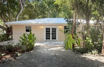 Single-story exterior featuring a metal roof, light-toned stucco facade, and double French doors, surrounded by mature trees and a gravel driveway