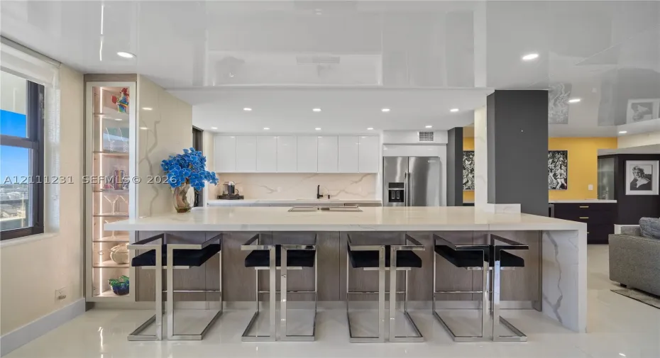 Kitchen featuring a waterfall-edge island with stone-finish countertop, high-gloss ceiling, integrated display cabinetry with interior lighting, sleek flat-panel cabinetry, and stainless steel refrigerator
