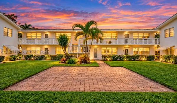 Two-story building exterior featuring a paver stone patio, manicured lawn, second-story balcony with decorative railing, and mature landscaping